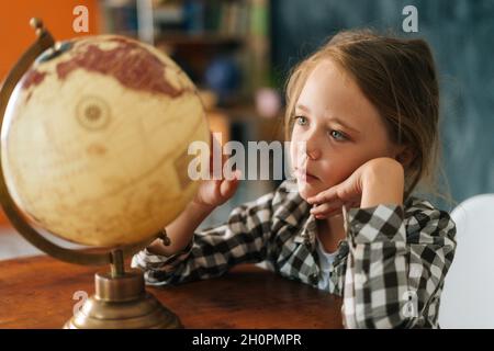 Primo piano di curiosa bambina che gira piccolo globo con interesse, guardando i paesi del mondo. Foto Stock