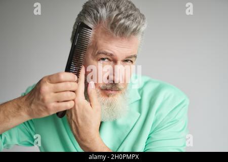 Ritratto di brutale uomo di mezza età con barba che guarda la fotocamera, spazzolando i capelli con il pettine, posando su sfondo bianco Foto Stock