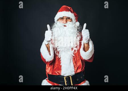 Uomo vestito come Babbo Natale con i pollici su sfondo nero. Concetto di Natale, Babbo Natale, regali, festa. Foto Stock