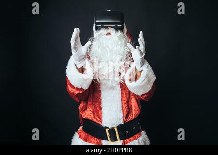 Uomo vestito come Babbo Natale indossando occhiali di realtà virtuale, su sfondo nero. Concetto di Natale, Babbo Natale, regali, festa. Foto Stock