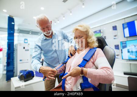 Coppia senior presso il negozio tecnologico. Donna che prova la tecnologia della realtà virtuale mentre l'uomo la aiuta a legare la cintura. Foto Stock