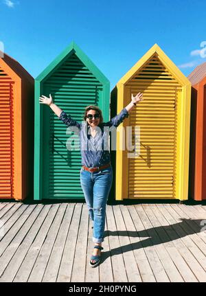 Felice sorgioiata giovane donna in abiti casual che alzano le mani sorridendo alla macchina fotografica mentre si trova contro colorate cabine in legno spiaggia in mare al sole Foto Stock