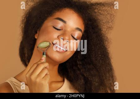 Studio girato di giovane donna afroamericana con occhi chiusi usando il rullo di giada facciale come trattamento della pelle mentre si alza isolato su sfondo marrone, Foto Stock