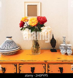 Bel bouquet di fiori di rosa freschi multicolore in vaso di vetro su tavolo giallo vintage con testa di buddha e vasi di ceramica antica di diverse dimensioni, Foto Stock