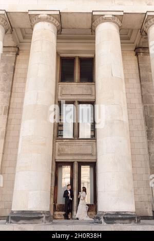 Giovane coppia, uomo e donna, sposa e sposo stanno in piedi su grandi passi di cemento tra colonne che tengono le mani, guardandosi l'un l'altro. Concetto di matrimonio Foto Stock