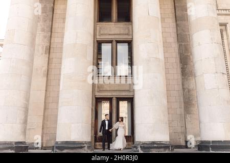 Giovane coppia, uomo e donna, sposa e sposo stanno in piedi su grandi passi di cemento tra colonne che tengono le mani, guardandosi l'un l'altro. Concetto di matrimonio Foto Stock