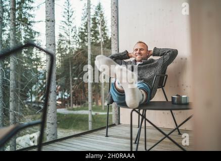 Ritratto di un uomo di mezza età sinceramente sorridente vestito cardigan aperto, jeans, e calze calde seduta in sedia sul balcone della casa foresta e godere del Foto Stock