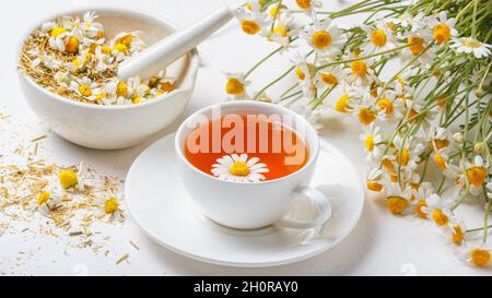 Tazza bianca di tè caldo della camomilla, un mortaio con boccioli di margherite secche e un bouquet di camomilla di Matricaria su fondo di cemento. Bevanda a base di erbe, sano li Foto Stock