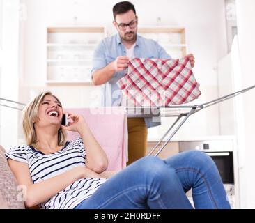 Bella giovane donna seduta sul divano, parlare sul telefono cellulare e ridere ad alto volume mentre l'uomo appende la lavanderia sul rack in background Foto Stock