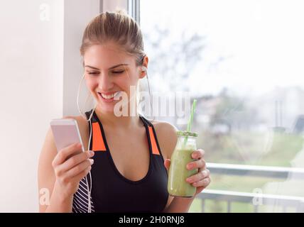 Giovane e graziosa donna in abbigliamento sportivo riposante dopo l'allenamento, bevendo frullati e ascoltando musica dallo smartphone. Concetto di stile di vita sano Foto Stock