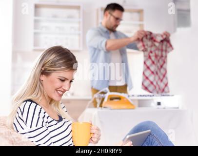 Bella giovane donna che riposa sul divano e guarda il tablet mentre l'uomo stira la sua camicia sullo sfondo. Marito che fa chores concetto Foto Stock