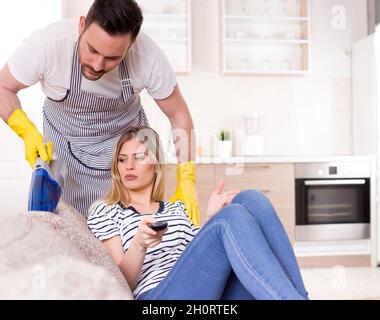 Giovane uomo con grembiule aspirapolvere divano mentre bella donna seduta e guardare la tv. La donna è infastidita dal marito che fa i lavori mentre sta riposando Foto Stock