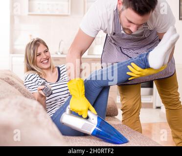 Giovane uomo con grembiule aspirapolvere divano con bella donna seduta e guardare la tv. Donna godendo mentre il marito fa i chores Foto Stock