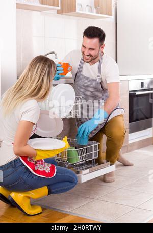 Giovane coppia felice scarico lavastoviglie con stoviglie pulite. Lavorando insieme e aiutarsi a vicenda nel lavoro domestico Foto Stock