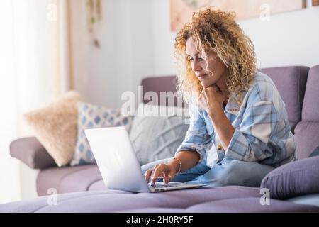 Sorridente hipster caucasico adulto sedersi rilassarsi sul divano utilizzando un moderno laptop che naviga senza limiti internet wireless, felice giovane donna freelance lavoro su comp Foto Stock