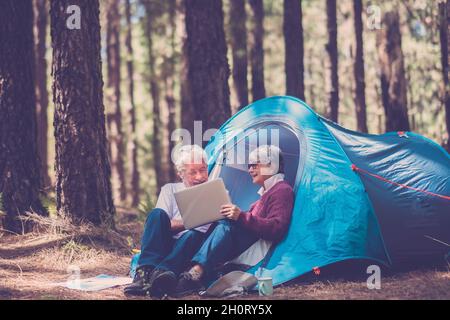 Le persone anziane attive amano il campeggio tenda all'aperto in un paesaggio forestale libero utilizzando un computer portatile per rimanere connessi - uomo e donna maturi in età outdoo Foto Stock