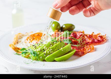 L'olio essenziale di cannabis CBD gocciola nel cibo. Colazione Keto. Uovo fritto con avocado e pancetta fritta in un piatto bianco. Foto Stock