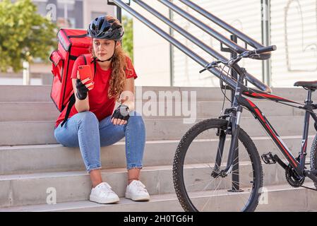 Una ragazza di consegna seduta sulle scale in attesa del prossimo ordine Foto Stock