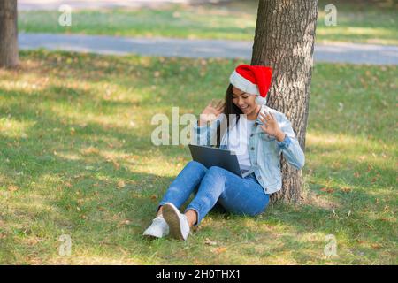 Giovane donna asiatica con un computer portatile nel parco. Si siede sull'erba a Santa Hat e comunica con i parenti alla vigilia di Natale. Video online Foto Stock