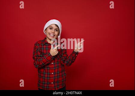 Adorabile ragazzo allegro preadolescente europeo, bello bambino in cappello di Santa e punti a scacchi maglia con le dita su uno spazio copia su sfondo rosso Foto Stock