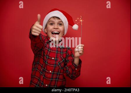 Fantastico ragazzo divertente bambino in Santa cappello e camicia a quadri tiene le luci del bengala, scintilla nelle sue mani, mostra il pollice in su, sorride guardando la macchina fotografica contro col Foto Stock