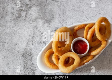 Anelli di cipolla in un piatto, con salsa di ketchup al pomodoro. Su sfondo concreto Foto Stock