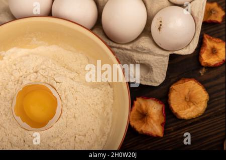Un uovo di pollo bianco rotto in una ciotola con farina di frumento, patatine secche di mela e diverse uova intere in un vassoio di cartone bianco, primo piano selettivo Foto Stock