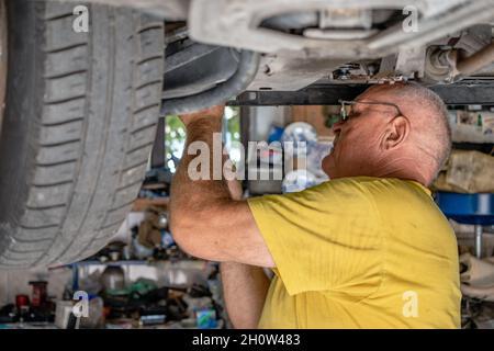 Meccanico auto anziano che controlla e ripara un'automobile sollevata nel suo garage. Controllo dei freni. Concetto di riparazione e meccanica delle auto vecchie. Foto Stock
