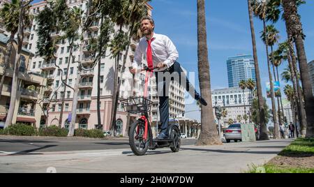 Felice direttore in abbigliamento formalwear ride stand-up e-scooter lungo il marciapiede, divertente Foto Stock