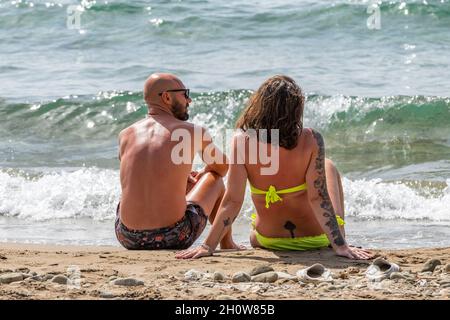 giovane coppia abbronzata e sana seduta insieme al sole sulla spiaggia in vacanza in grecia. giovane coppia seduta sulla spiaggia a zante. Foto Stock