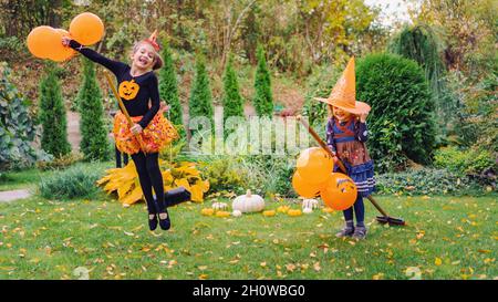 I bambini in cappelli di strega ridono e giocano con le scopa. Le streghe piccole imparano a volare i bromoomsticks con i palloncini d'arancia ad una festa all'aperto di Halloween nel f Foto Stock