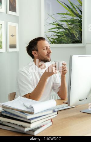 Un uomo d'affari casual si siede al posto di lavoro del computer bere caffè, rilassarsi dal lavoro con i documenti finanziari Foto Stock