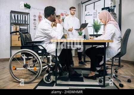 Medico africano che utilizza la sedia a rotelle seduto con i suoi colleghi multirazziali a tavola e ascoltando il docente caucasico durante la riunione. Seminario medico di specialisti competenti. Foto Stock