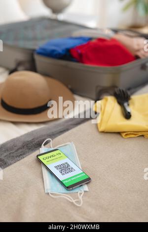 Smartphone con comodo passaporto, maschera facciale e valigia appoggiata sul letto Foto Stock