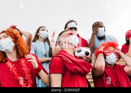 Frustrato gruppo di tifosi di calcio multirazziale in maschera protettiva che guarda la loro squadra di calcio perdere - Focus su uomo nel centro Foto Stock