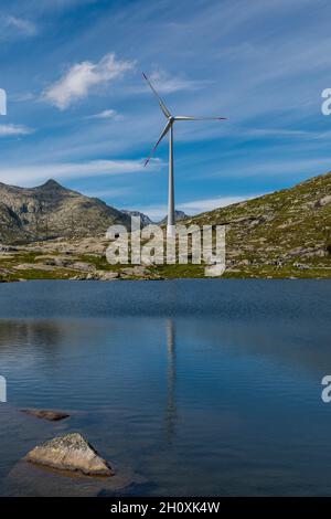 La turbina eolica in cima al valico del Gottardo nelle Alpi svizzere si riflette nelle acque del lago. Spazio copia per le energie rinnovabili. Foto Stock