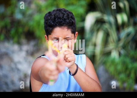 Ragazzo giovane e giocoso che punta a una semplice catapulta fatta in casa alla fotocamera che squinting con un occhio per garantire la precisione all'aperto in giardino Foto Stock