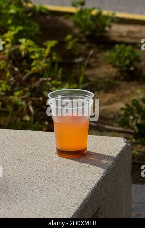 Primo piano di una tazza di plastica con una bevanda arancione che qualcuno ha lasciato su una panchina in un parco pubblico in una città. Problemi delle bottiglie per giovani nelle città, garba Foto Stock