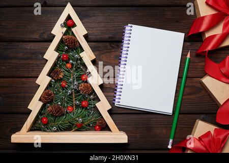 Albero di natale in legno con rami di abete all'interno, scatole regalo con nastri rossi e un blocco note vuoto su sfondo di legno Foto Stock