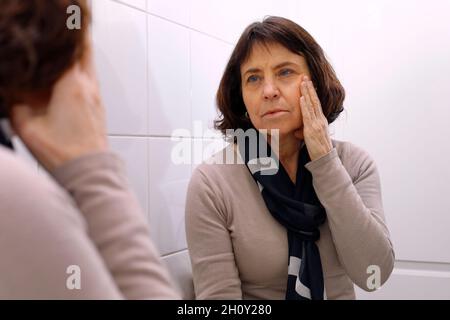 Una donna matura che guarda nello specchio il suo volto Foto Stock