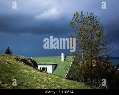 HAFNARFJORDUR, ISLANDA - 31 maggio 2021: Una casa in stile scandinavo con tetto verde e camino, nascosto dietro collina verde. Cielo scuro, tempestoso. Foto Stock