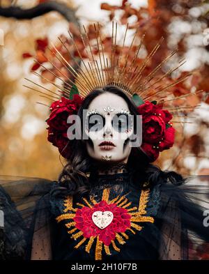 Ritratto di giovane donna con trucco del cranio dello zucchero e rose rosse vestite in costume nero della morte come Santa Muerte sullo sfondo delle foglie autunnali dentro Foto Stock