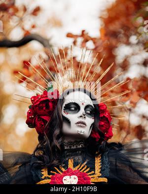 Ritratto di giovane donna con trucco del cranio dello zucchero e rose rosse vestite in costume nero della morte come Santa Muerte sullo sfondo delle foglie autunnali dentro Foto Stock