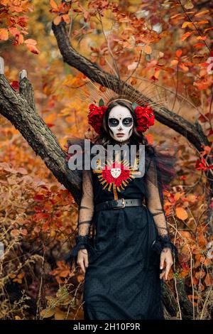 Giovane donna con trucco del cranio dello zucchero e rose rosse vestite in costume nero della morte come Santa Muerte è sullo sfondo delle foglie autunnali nella foresta. Foto Stock