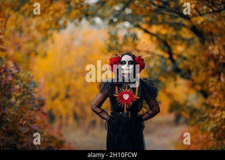 Giovane donna con trucco del cranio dello zucchero e rose rosse vestite in costume nero della morte come Santa Muerte è sullo sfondo della foresta autunnale. Giorno del Foto Stock