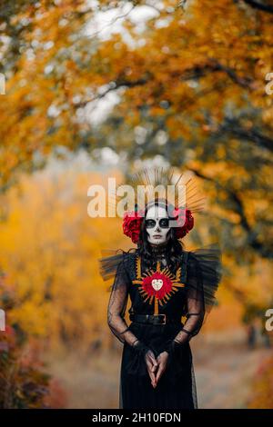 Giovane donna con trucco del cranio dello zucchero e rose rosse vestite in costume nero della morte come Santa Muerte è sullo sfondo della foresta autunnale. Giorno del Foto Stock