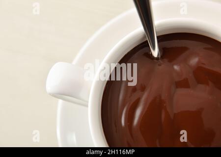 Cioccolato caldo in tazza di ceramica bianca con cucchiaio di metallo su un piatto su tavola di legno bianco. Vista dall'alto. Composizione orizzontale. Foto Stock