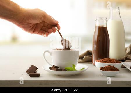 Mescolare a mano il cioccolato caldo appena preparato sul banco da cucina con porzioni e cioccolato in polvere e bottiglie con latte e cioccolato liquido sul kitch Foto Stock