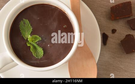 Macro di cioccolato caldo in tazza di ceramica bianca su un piatto con cucchiaio di legno e foglie di menta e pezzi di cioccolato su tavola di legno. Vista dall'alto. Compo. Orizzontale Foto Stock