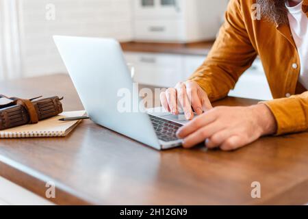 L'uomo usa un notebook per l'educazione o lo shopping online sul lavoro. Primo piano mani maschili giovane uomo che lavora da casa al computer portatile in cucina a casa. Freelance Foto Stock